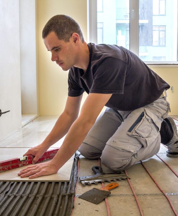 Young worker tiler installing ceramic tiles using lever on cement floor with heating red electrical cable wire system. Home improvement, renovation and construction, comfortable warm home concept.